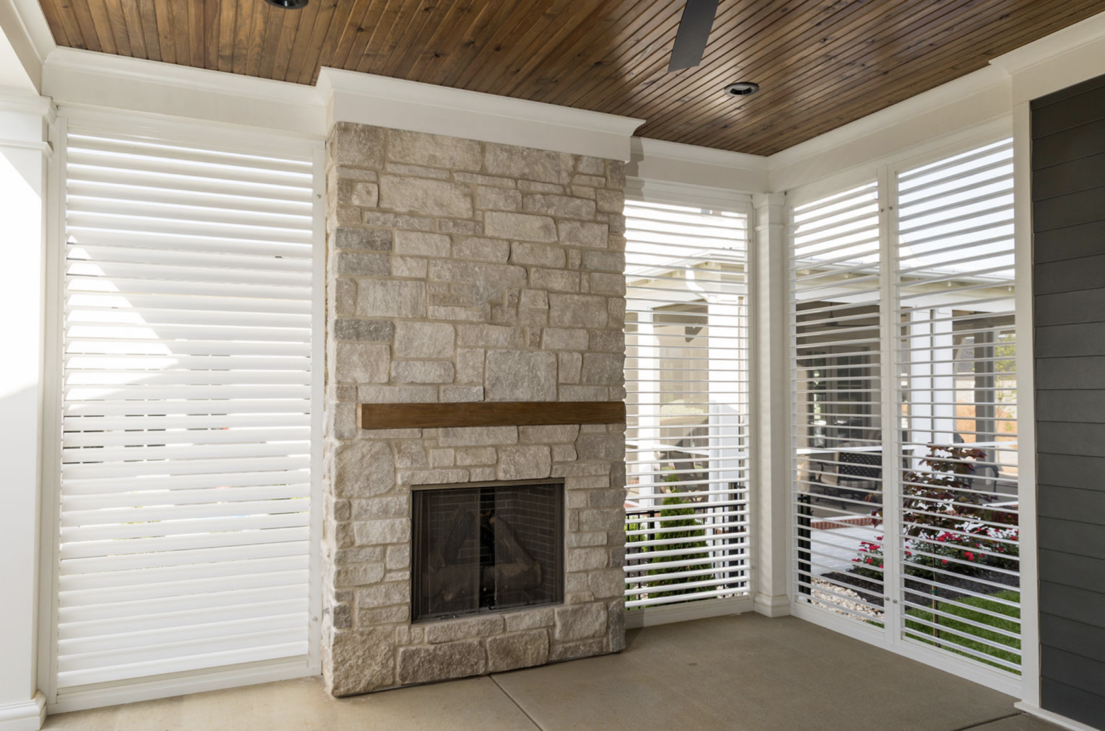 Covered outdoor patio with a natural stone fireplace, wooden ceiling, and white plantation shutters providing partial views of a landscaped backyard.