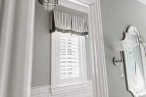 Elegant Louisville bathroom window with floral valance, white blinds, chandelier, and granite countertop by Draped in Style experts.